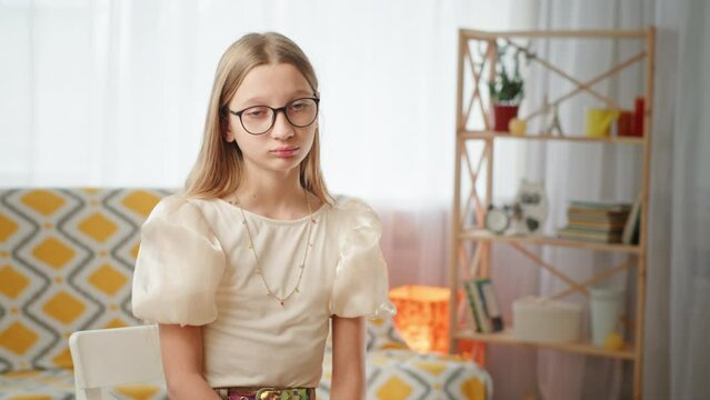 Upset girl looking at camera. Sad girl in stylish blouse and glasses looking at camera and gasping while sitting on chair near sofa in living room at home.