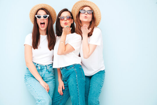 Three young beautiful smiling hipster female in trendy same summer white t-shirt and jeans clothes. carefree women posing near light blue wall in studio. Cheerful and positive models give air kiss