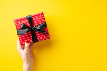 Black friday shopping concept. First person top view photo of female hand giving red giftbox with ribbon bow on isolated yellow background with copyspace