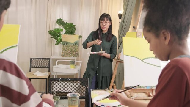 A female Asian teacher teaches and demonstrates to the children on acrylic color picture painting on canvas in art classroom, creatively learning with skill at the elementary school studio education.