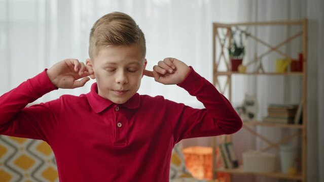 Boy rolls eyes and covers ears with fingers shaking head. Irritated junior schoolboy in crimson shirt rolls eyes and covers ears with fingers shaking head. Child purses lips and closes eyes breathing