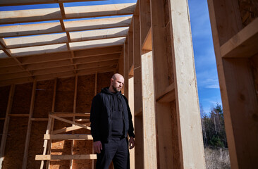 Fototapeta premium Male engineer building wooden frame house. Bald man standing on construction site, inspecting quality of work on sunny day.