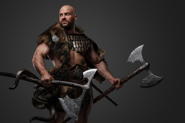Studio shot of handsome ancient viking dressed in armor and fur holding two axes.