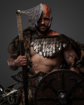 Shot of muscular viking with bloody face holding axe and shield against grey background.