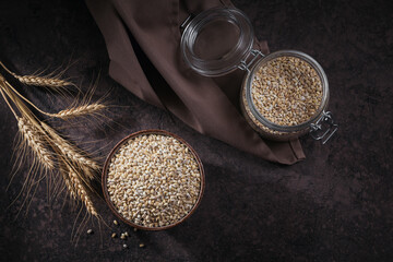 Organic uncooked dried barley cereal grain in a bowl with ears of wheat on white wooden background....