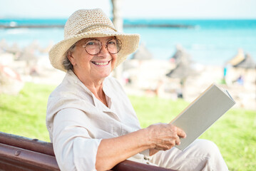 Beautiful smiling elderly woman sitting on a bench in front of the sea relaxing with a book -...