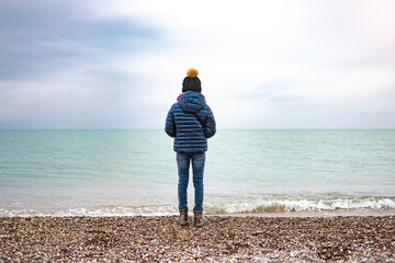 The girl stands with her back to the sea in a warm jacket and knitted hat.