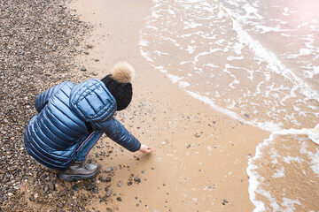 Obraz premium A girl in a warm jacket and knit cap touching the water in the sea while standing on the shore.