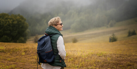 Happy senior elderly woman traveler with a backpack standing near the foggy forest. Active...