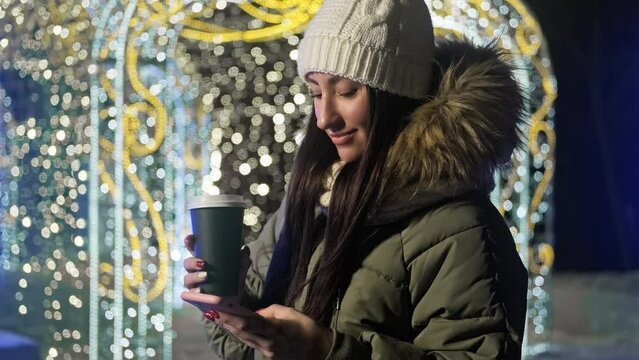 Young woman using her phone via texting with hot coffee in hands. Female in knitted hat with xmas lights at background. Christmas concept. Winter evening. 4K, UHD