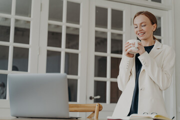 Smiling female employee standing at office desk enjoying break with coffee cup during work at laptop