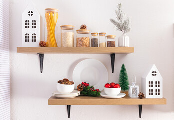 Fototapeta na wymiar Front view of open kitchen shelves with eco jars for bulk products and Christmas decorations. cardboard white houses, natural cones.