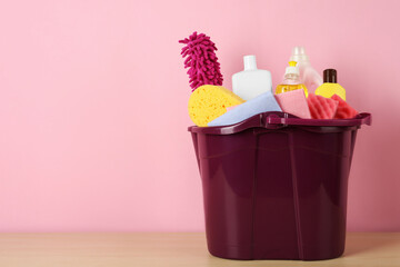 Fototapeta na wymiar Bucket with different cleaning supplies on wooden floor near pink wall. Space for text