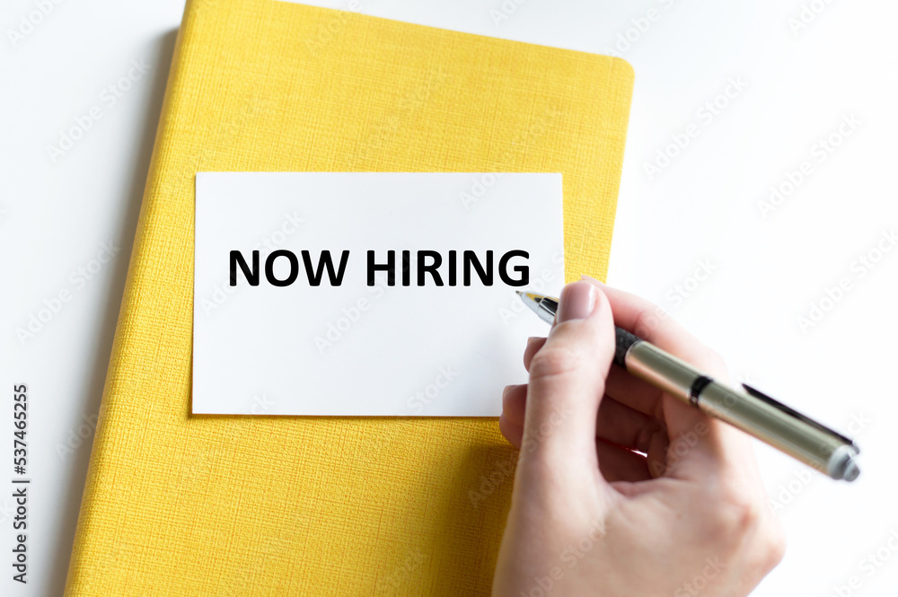 Poster now hiring text on a card against the background of a yellow notebook on a table of a person's hand