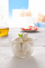 morsels of mozzarella in a glass bowl, with slices of tomato and olive oil placed on a white wooden table