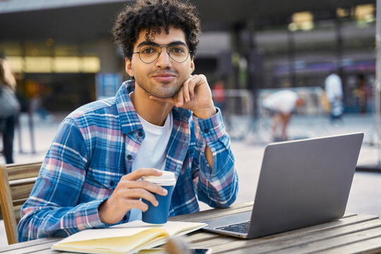 Portrait of young man freelancer using laptop computer working online holding cup of coffee. Middle eastern student studying looking at the camera, online education concept  