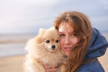 portrait of beautiful happy positive girl, young woman walking with her Pomeranian Spitz dog, holding puppy on hands, smiling, having fun outdoors at beach