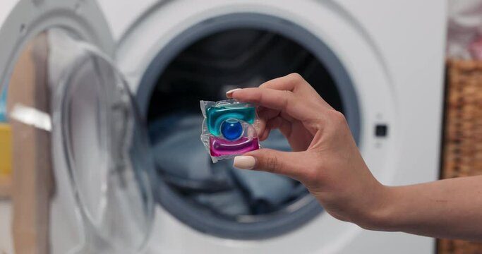 Close-up of hands holding a laundry capsule and dropping it into the drum of a washing machine with clothes ready to be turned on.
