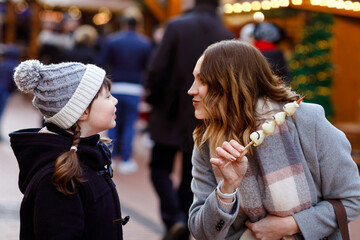 Mother and daughter eating white chocolate covered fruits and strawberry on skewer on traditional German Christmas market. Happy girl and woman on traditional family market in Germany during snowy day