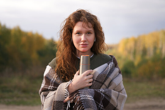 Portrait of beautiful girl, young happy woman is drinking tea or coffee, hot beverage from thermo cup, thermos bottle, covering herself in plaid, cozy blanket in natural golden autumn park or forest