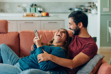 happy couple hugging using cellphone together lying on sofa at home - laughing joyful man and woman with smartphone at home - young married with mobile phone relaxing on couch