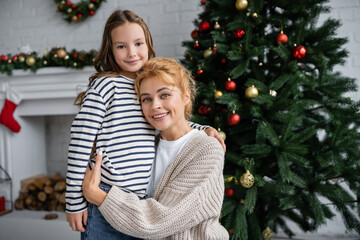 Obraz na płótnie Canvas Smiling mom in knitted cardigan hugging daughter near christmas tree at home