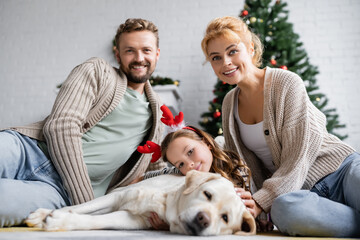Positive parents looking at camera near daughter with christmas headband and labrador on floor at home