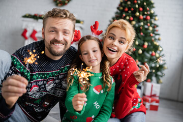Positive family in warm sweaters holding sparklers during new year celebration at home