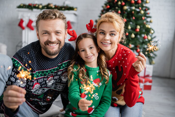 Cheerful family in christmas sweaters holding sparklers and looking at camera at home