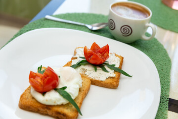 Breakfast with cup of coffee and whole wheat toasted bread with poached egg, tomato, arugula and cheese on white plate.
