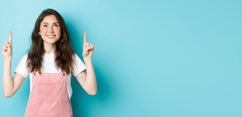 Fototapeta na wymiar Young cheerful girl with glamour make up, wearing pink summer dungarees, pointing fingers up and smiling, looking at promo offer on top, blue background