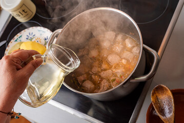 Woman's hand with a jug adding white wine to a pot with stewed meat in the kitchen at home.