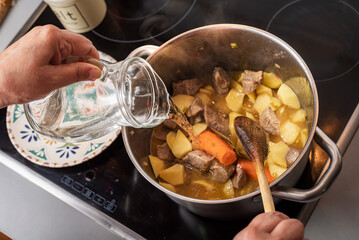 Woman's hand with a jug adding water to a pot with stewed meat and potatoes in the kitchen at home.