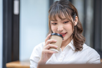  Smiling business Asian woman sitting at workplace office desk, holding a cup of coffee, she is relaxing and looking on laptop, report real estate investment data, Financial and tax systems concept.