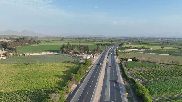 Aerial drone shot of vehicles crossing four lane Panamericana Norte highway surrounded by mountains in Peru through farming fields on both corners on a sunny day