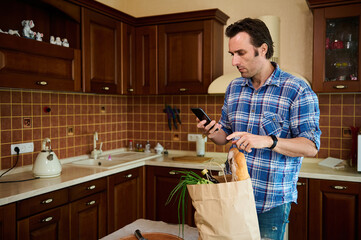 Healthy shopping at the grocery store. Food delivery. Handsome young Caucasian man in blue plaid shirt, checking expenses while returning home after grocery shopping and unpacking healthy food bag