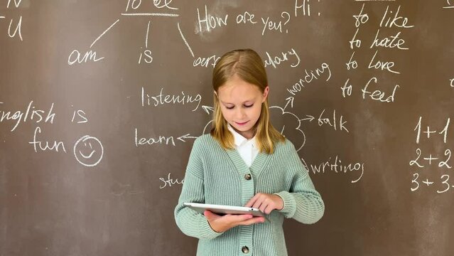 little girl using a digital tablet standing at the painted blackboard in the classroom. child fingers touching the screen. Preschool kid hold pad computer surfing internet for education indoors. 