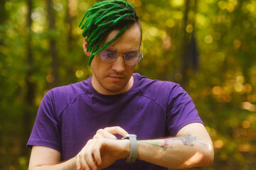 Man using smart watch in park. Male in violet t shirt with glasses and green deadlocks checking...