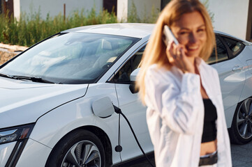 Fototapeta na wymiar Holding phone and talking by it. Young woman in white clothes is with her electric car at daytime
