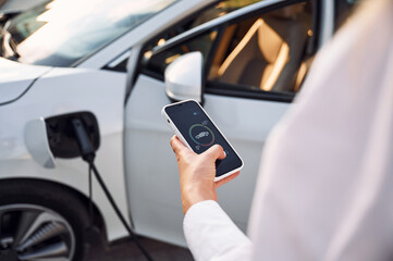 Smartphone monitoring of charging. Young woman in white clothes is with her electric car at daytime