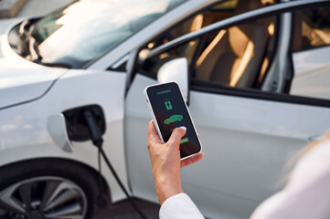 Smartphone monitoring of charging. Young woman in white clothes is with her electric car at daytime
