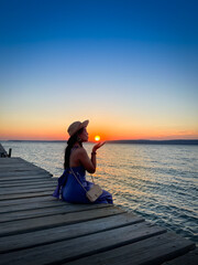 Abstract lifestyle, Traveler woman with summer vacations on twilight by sea her sitting on bridge lonely, holds the sun in hand and pray. Aegean Sea Beach, Canakkale, Turkye. Blank space for text.