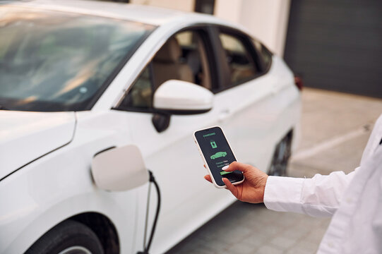 Near Garage. Close Up View. Woman Charging Her Electric Car And Monitoring Process On Smartphone