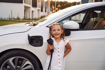 Little girl standing near electric car with charging cable in hands