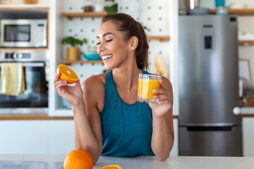 Beautiful young woman drinking fresh orange juice in kitchen. Healthy diet. Happy young woman with glass of juice and orange at table in kitchen. - obrazy, fototapety, plakaty