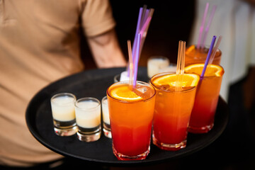 The waiter holds a tray with Multicolored alcoholic and non-alcoholic cocktails with ice and straws