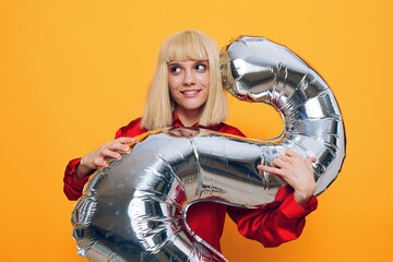 horizontal photo on a yellow background of a happy, joyful woman standing in a red shirt with a balloon in the form of the number two in silver color