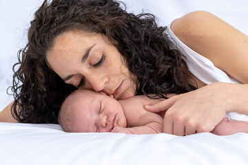 mother hugs and tenderly looks at her newborn baby while she sleeps and rests on the bed in the room. White background. family and affection concept