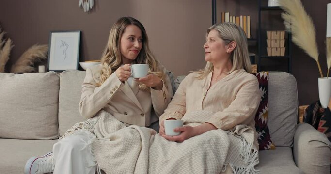 Mother and daughter spend an afternoon together on the living room couch. The women drink coffee and tea and talk, joke around.