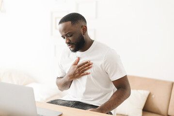 A black man feels pain in his heart while sitting at home and working on a laptop. Heart problems...